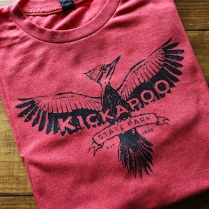 May include: Red t-shirt with a black graphic of a bird with outstretched wings and the words "KICKAPOO STATE PARK EST. 1939" in a banner. The shirt is folded on a wooden surface.