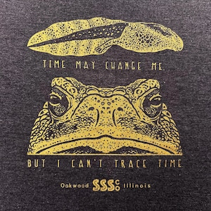 May include: Dark gray t-shirt with a gold illustration of a tadpole above a frog's face. The text reads "TIME MAY CHANGE ME. BUT I CAN'T TRACE TIME." Below the text is "Oakwood SSSS Illinois."