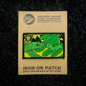 May include: Iron-on patch featuring a green snake and a green caterpillar on a yellow background with green leaves. The patch has a black border and the text "SSS CO" in a white circle in the top left corner. The text "IRON-ON PATCH" and "SMILINGSNAKE.ETSY.COM" are printed below the patch.