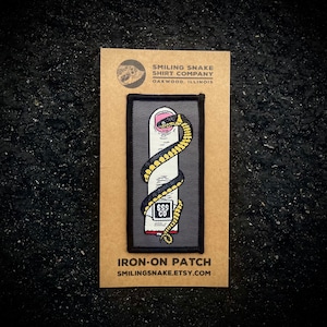 May include: Iron-on patch featuring a black snake with yellow stripes wrapped around a finger. The patch has a gray background and the text "Smiling Snake Shirt Company" and "Iron-on Patch" are printed on the card.