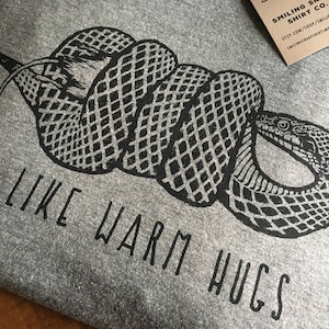 May include: A gray t-shirt with a black graphic of a coiled snake. The text "I LIKE WARM HUGS" is printed below the snake. A small brown tag with white text is in the upper right corner.