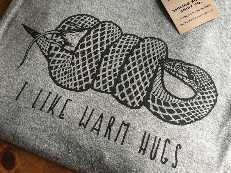May include: A gray t-shirt with a black graphic of a coiled snake. The text "I LIKE WARM HUGS" is printed below the snake. A small brown tag with white text is in the upper right corner.