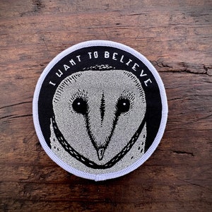 May include: A black and white embroidered patch with a white owl face and the text "I WANT TO BELIEVE" in a white circle with a white border.