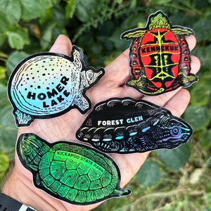 May include: A collection of four turtle-shaped stickers. One sticker says "HOMER LAKE" in blue and white. Another says "KENNEKUK" in red and yellow. The third says "FOREST GLEN" in white. The last says "KICKAPOO STATE PARK" in green.