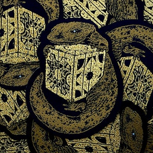 May include: A close-up of a gold-colored embroidered patch featuring a detailed design of a creature holding a cube. The creature appears to be a snake or lizard with a textured, scaly body. The cube is intricately patterned with geometric shapes and lines.