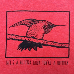 May include: Red t-shirt featuring a black illustration of a hummingbird perched on a branch within a black rectangular frame. The text "LIFE'S A BUMMER WHEN YOU'RE A HUMMER" is printed below the frame in black.