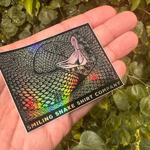 May include: A holographic sticker featuring a detailed illustration of a snake with its mouth open. The sticker has a rainbow sheen and the text "SMILING SNAKE SHIRT COMPANY" is printed at the bottom.