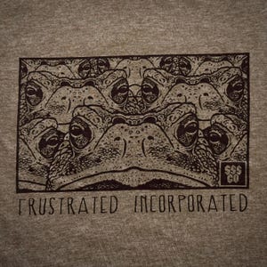 May include: A tan t-shirt featuring a rectangular graphic of multiple toad faces in a repeating pattern. The text "FRUSTRATED INCORPORATED" is printed below the graphic in a dark brown color. A small logo is in the bottom right corner.