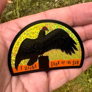 May include: A black sticker with a vulture illustration. The vulture is perched on a black base, set against a yellow and orange background. The text "I wanna soak up the sun" is printed on the sticker.