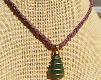 May include: A green glass pendant wrapped in gold wire hangs from a brown cord necklace. The necklace is displayed on a tan fabric background.
