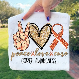 May include: White sweatshirt with a graphic design featuring a peace sign, heart, and ribbon, with the words "peace, love, cure" and "COPD AWARENESS". The peace sign and ribbon have a pattern of orange ribbons.