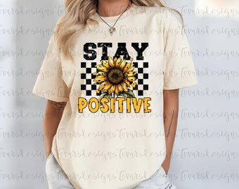 May include: A cream-coloured t-shirt with a sunflower graphic and the words "STAY POSITIVE" in black and yellow. The design includes a black and white checkered pattern behind the sunflower.