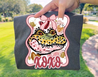 May include: Dark gray t-shirt featuring a colorful alligator design with a pink bow and the text "xoxo." The alligator has yellow, green, and pink accents. The design is framed with a pink border and gold glitter.