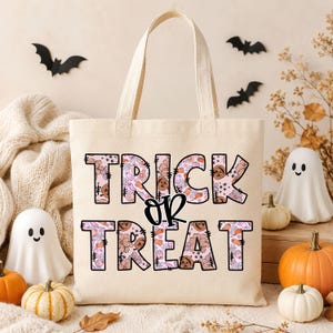May include: A cream-colored canvas tote bag featuring the text "TRICK OR TREAT" in large block letters. The letters are filled with a repeating pattern of small orange pumpkins, pink ghosts, and spiderwebs on a light pink background.