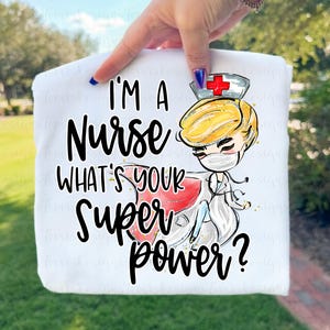 Könnte beinhalten: Ein weißes T-Shirt mit einer grafischen Illustration einer Pflegekraft mit blonden Haaren, die einen Mundschutz und eine Haube mit rotem Kreuz trägt. Der Text lautet I'M A Nurse WHAT'S YOUR SUPER power? in schwarzer Schrift.