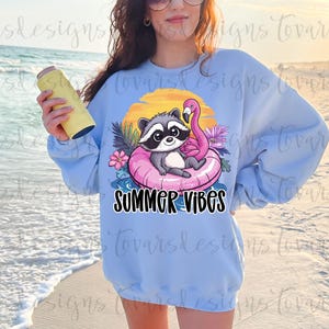 May include: Light blue sweatshirt with a graphic of a raccoon in a pink inflatable ring with a flamingo, flowers, and the text "SUMMER VIBES". A yellow drink holder is held in the hand. The background is a beach scene.