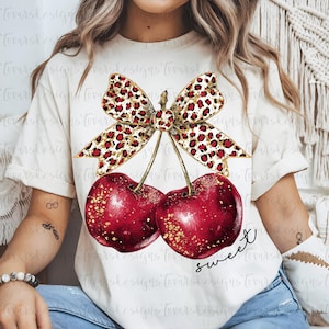 Puede incluir: Camiseta color crema con un gráfico de dos cerezas rojas con detalles de purpurina dorada y un lazo con estampado de leopardo. La palabra "sweet" está escrita en cursiva debajo de las cerezas.