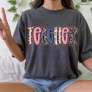 K&ouml;nnte beinhalten: Dunkelgraues T-Shirt mit dem Wort "TEACHER" in gro&szlig;en, bunten Buchstaben. Die Buchstaben sind goldfarben umrandet und mit rosa und blauen Mustern versehen. Das Shirt ist im l&auml;ssigen Stil.