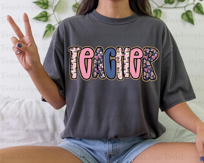 K&ouml;nnte beinhalten: Dunkelgraues T-Shirt mit dem Wort "TEACHER" in gro&szlig;en, bunten Buchstaben. Die Buchstaben sind goldfarben umrandet und mit rosa und blauen Mustern versehen. Das Shirt ist im l&auml;ssigen Stil.
