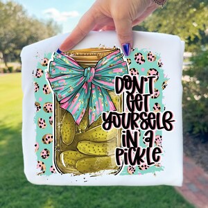 Peut inclure: T-shirt blanc avec un motif de bocal de cornichons orné d'un nœud turquoise et rose. Le texte "DON'T GET YOURSELF IN A PICKLE" est imprimé en noir et blanc. Le motif comprend un fond turquoise avec des taches léopard roses et noires.