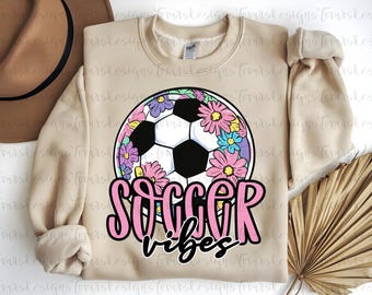 May include: Beige sweatshirt with a soccer ball graphic surrounded by colorful flowers. The words "Soccer Vibes" are written in pink and black script. A casual, sporty design.