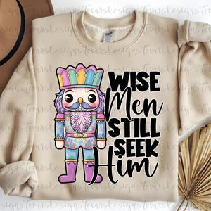 May include: Beige sweatshirt featuring a colorful nutcracker design with the text "WISE Men STILL SEEK Him" in black. The nutcracker has a pink beard, a crown, and a detailed uniform. A brown hat and dried palm leaves are in the background.