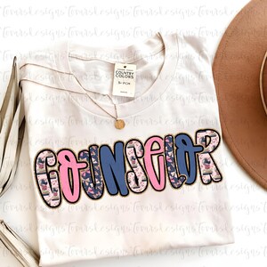 K&ouml;nnte beinhalten: Cremefarbenes T-Shirt mit dem Wort "COUNSELOR" in einer Blasen-Schriftart. Die Buchstaben sind mit goldenem Glitzer umrandet, mit rosa und blauen Farben und einem Muster aus B&uuml;chern und Herzen. Eine goldene Halskette und ein brauner Hut sind ebenfalls zu sehen.