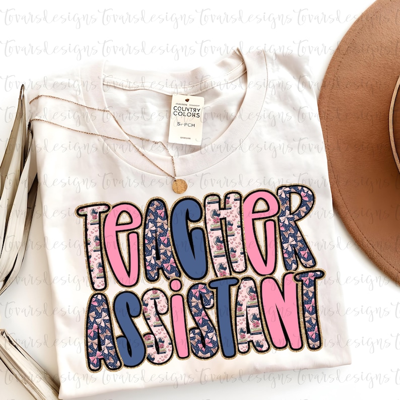 K&ouml;nnte beinhalten: Ein cremefarbenes T-Shirt mit den Worten "TEACHER ASSISTANT" in einer Blasen-Schriftart. Die Buchstaben sind schwarz umrandet und mit rosa, blauen und gemusterten Designs gef&uuml;llt. Eine goldene Halskette und ein brauner Hut sind ebenfalls sichtbar.