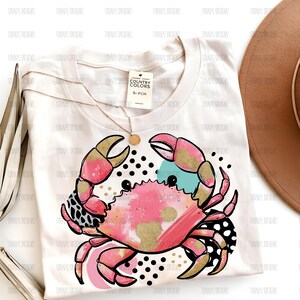 May include: A white t-shirt featuring a graphic of a crab with pink, gold, and teal watercolor-style textures, accented by black polka dots and a leopard print pattern. A small tag labeled Country Colors S-PCH is attached to the neckline.