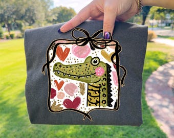 May include: Dark gray t-shirt featuring a colorful alligator design. The alligator is green with pink cheeks and is surrounded by hearts and gold accents. The design is framed with a black border and a bow.