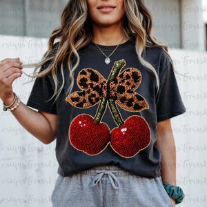 以下が含まれることがあります： チャコールグレーの半袖Tシャツで、緑の茎がついた2つの赤いサクランボと、茶色のヒョウ柄のリボンが大きく描かれています。グラフィックはビーズのような質感です。Tシャツにはグレーのドローストリング付きスウェットパンツが合わせられています。
