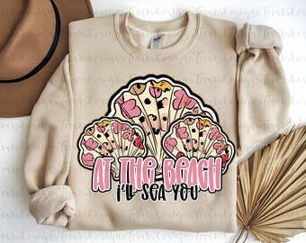 May include: A beige sweatshirt with a seashell graphic. The graphic features three stylized seashells with pink floral patterns and the text "AT THE BEACH I'LL SEA YOU" in pink and black.