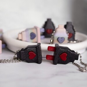 May include: Two black plastic keychains shaped like electrical plugs with red heart accents. The keychains are connected by a silver chain.