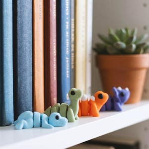 May include: A row of four small, articulated dinosaur figurines in light blue, light green, orange, and purple. Each dinosaur has a rounded head, large black eyes, and a segmented body structure, positioned in a line on a white shelf surface.