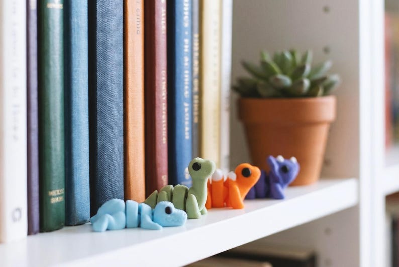May include: A row of four small, articulated dinosaur figurines in light blue, light green, orange, and purple. Each dinosaur has a rounded head, large black eyes, and a segmented body structure, positioned in a line on a white shelf surface.