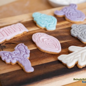 May include: Assortment of decorated cookies on a wooden cutting board. Cookies include shapes like a dragon, airplane, robot, and a cookie with the word "FEMIN" in pink icing. Other cookies are light blue, pink, and purple.