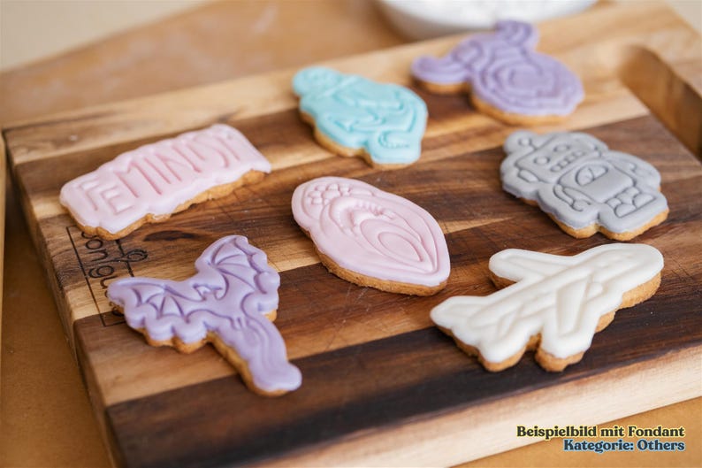 May include: Assortment of decorated cookies on a wooden cutting board. Cookies include shapes like a dragon, airplane, robot, and a cookie with the word "FEMIN" in pink icing. Other cookies are light blue, pink, and purple.