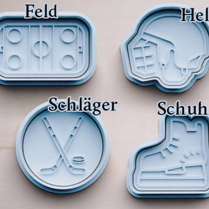 May include: Four blue plastic cookie cutters shaped like hockey equipment. The cutters are labeled "Feld", "Helm", "Schl&auml;ger", and "Schuhe".