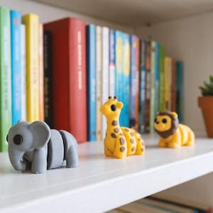 May include: Three small articulated animal figurines made of matte material, including a gray elephant, a yellow giraffe with brown spots, and a yellow lion with a brown mane, arranged in a row on a white shelf in front of a bookshelf.