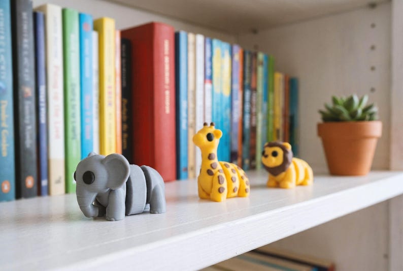 May include: Three small articulated animal figurines made of matte material, including a gray elephant, a yellow giraffe with brown spots, and a yellow lion with a brown mane, arranged in a row on a white shelf in front of a bookshelf.