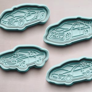 May include: Four light blue plastic cookie cutters shaped like different cars. The cutters are on a light brown surface.