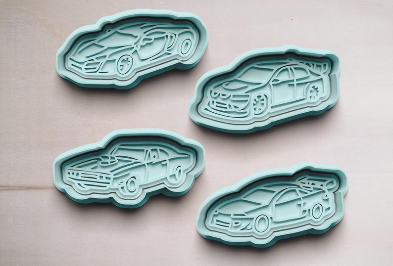 May include: Four light blue plastic cookie cutters shaped like different cars. The cutters are on a light brown surface.