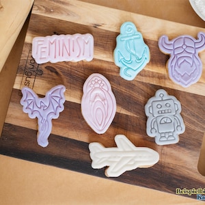 May include: A wooden cutting board displays various cookie cutters in the shapes of an anchor, robot, airplane, bat, Viking, and a design with the word "FEMINISM". The cookies are in pastel colors.