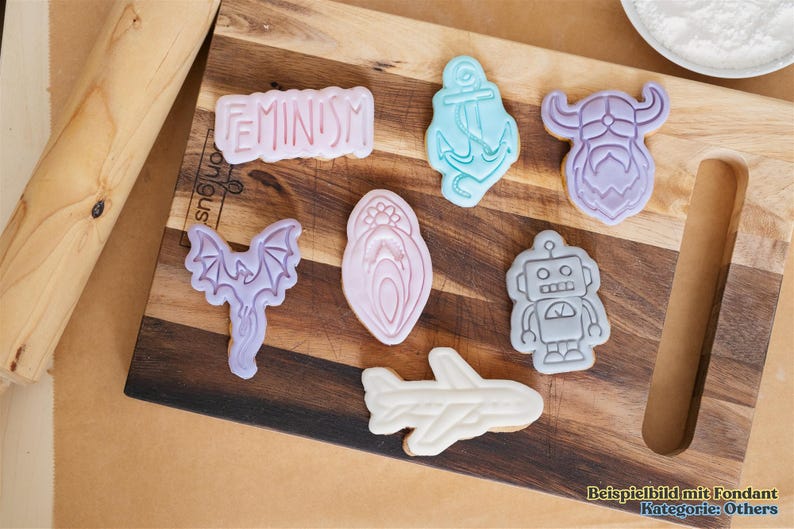 May include: A wooden cutting board displays various cookie cutters in the shapes of an anchor, robot, airplane, bat, Viking, and a design with the word "FEMINISM". The cookies are in pastel colors.