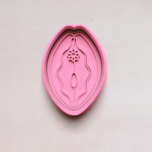 May include: A pink cookie cutter in the shape of a stylized vulva. The cutter has a layered design with a floral element at the top. The object is set against a light-colored background.