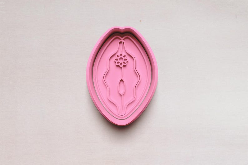 May include: A pink cookie cutter in the shape of a stylized vulva. The cutter has a layered design with a floral element at the top. The object is set against a light-colored background.