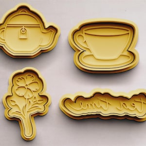 Könnte beinhalten: Vier gelbe Ausstecher, einer in Form einer Teekanne, einer in Form einer Teetasse und Untertasse, einer in Form einer Blume und einer mit dem Text "Tea Time" in einer Schreibschrift.