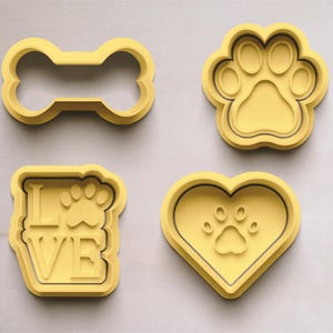 Puede incluir: Cuatro cortadores de galletas de plástico amarillo en forma de hueso, una huella de pata, un corazón con una huella de pata dentro y un rectángulo con la palabra "LOVE" y una huella de pata dentro.