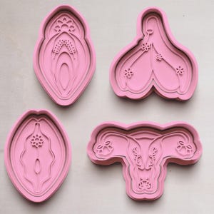 May include: Four pink cookie cutters in various shapes, including a stylized vulva and uterus. The designs feature detailed floral and layered elements, set against a light wooden background.