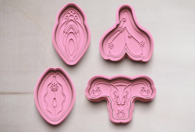 May include: Four pink cookie cutters in various shapes, including a stylized vulva and uterus. The designs feature detailed floral and layered elements, set against a light wooden background.